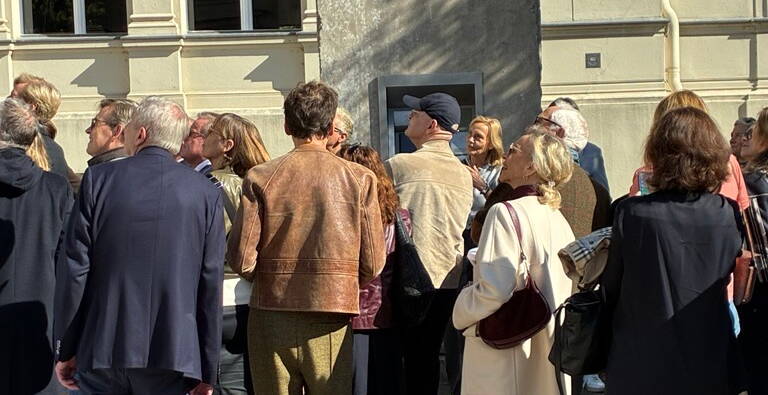 Eine Gruppe von Menschen versammelt sich vor einem Gebäude mit einer grauen Wandinstallation in der Sonne. (Text KI-generiert)