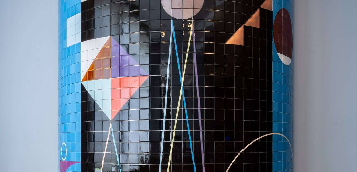 Ein zylindrisches Mosaik mit geometrischen Formen und bunten Kacheln schmückt einen Abschnitt einer Wand.