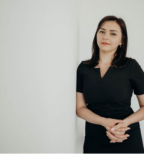 A woman in a black dress stands confidently against a white wall, conveying a sense of professionalism and poise.