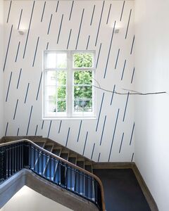 A staircase with a black railing ascends beside a window and a wall adorned with blue diagonal lines. | © VG Bild-Kunst