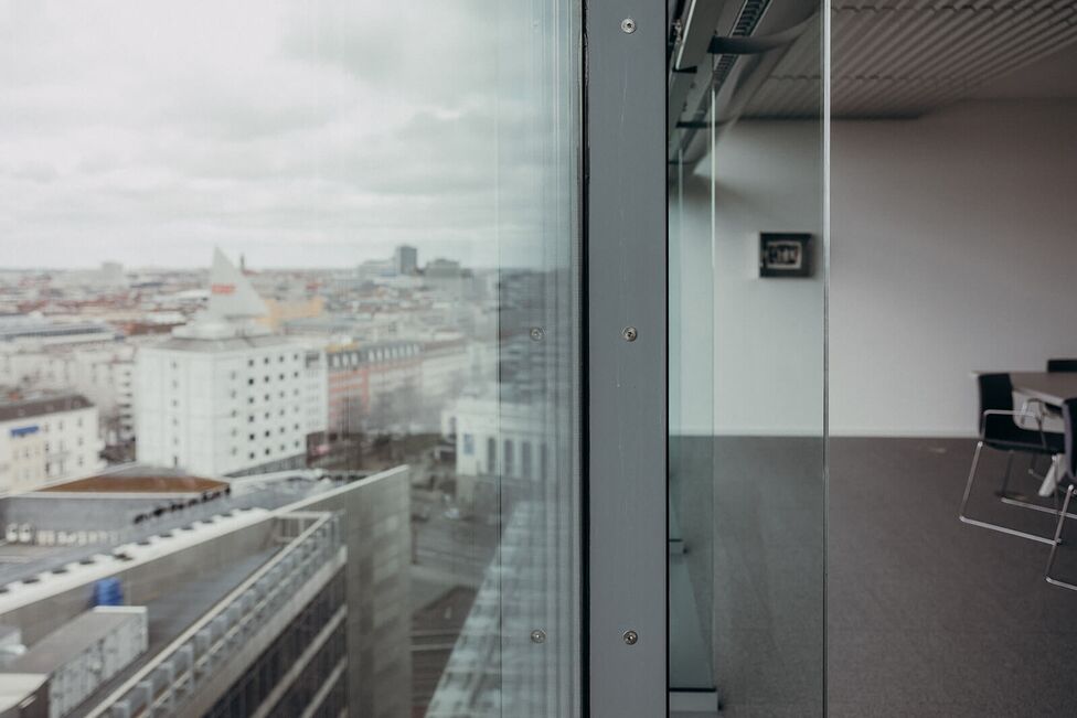 Blick aus einem modernen Büro mit Glaswand auf eine städtische Skyline unter wolkenverhangenem Himmel.