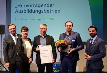 Eine Gruppe von fünf Personen posiert auf einer Bühne, während eine Person eine Auszeichnung und Blumen hält.