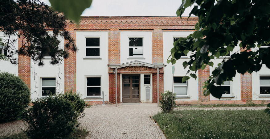 A brick building with a "Kontor" sign is framed by lush trees, creating a serene atmosphere.