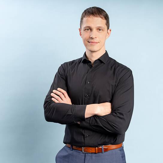 A confident man in a black shirt stands with arms crossed against a light blue background.