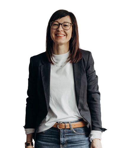A smiling woman in glasses and a blazer stands confidently against a white background.