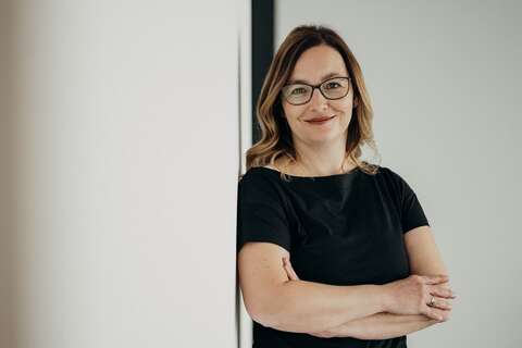 A woman in glasses and a black dress stands confidently with arms crossed, leaning against a white wall.