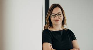 Eine lächelnde Frau mit Brille lehnt entspannt an einer weißen Wand und trägt ein schwarzes Kleid.