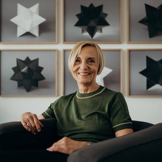 Eine lächelnde Person sitzt vor einer Wand mit geometrischen Kunstwerken in einem modernen Raum.