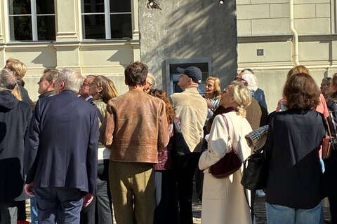 Eine Gruppe von Menschen versammelt sich vor einem Gebäude mit einer grauen Wandinstallation in der Sonne. (Text KI-generiert)