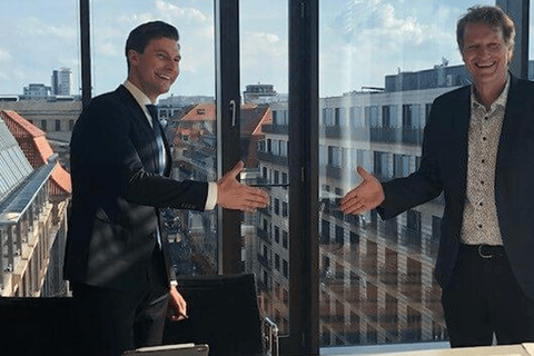 Zwei Männer in Anzügen stehen in einem modernen Büro vor einer Glasfront und bereiten sich auf einen Händedruck vor.