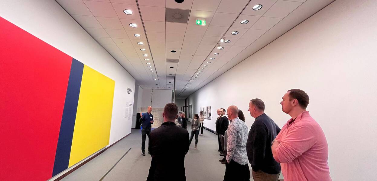 Mehrere Personen betrachten ein großes, farbenfrohes Wandgemälde in einer modernen Kunstgalerie.