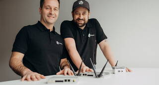Zwei Männer in schwarzen Poloshirts stehen lächelnd an einem Tisch mit mehreren technischen Geräten.