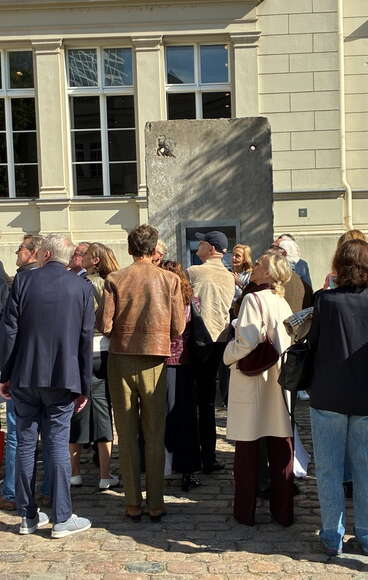 A crowd gathers outside a building, admiring an art installation against a wall on a sunny day. (Text KI-generiert)