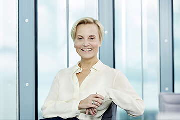 A person in a white shirt smiles confidently, sitting in front of large glass windows, conveying a sense of news professionalism.