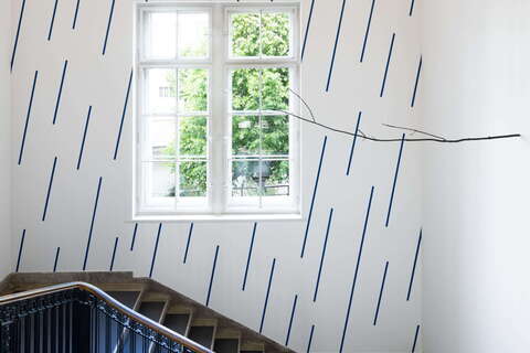 A staircase with a black railing ascends beside a window and a wall adorned with blue diagonal lines. | © VG Bild-Kunst
