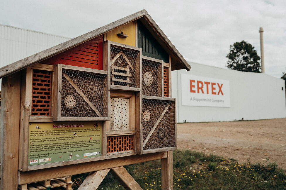A wooden insect hotel stands in front of a building with the sign "ERTEX, A Peppermint Company," surrounded by greenery.