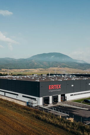 Ein modernes Industriegebäude von Ertex steht vor einer malerischen Berglandschaft, mit Autos auf dem Parkplatz.