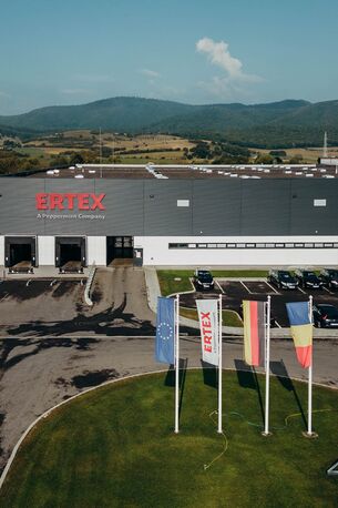Ein modernes Gebäude von Ertex International mit geparkten Autos und wehenden Flaggen vor einer malerischen Bergkulisse.