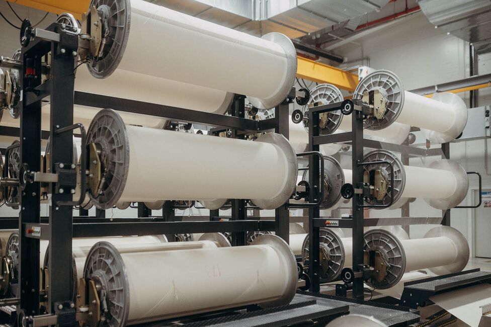 Large industrial spools of white fabric are neatly arranged on metal racks in a textile production facility.