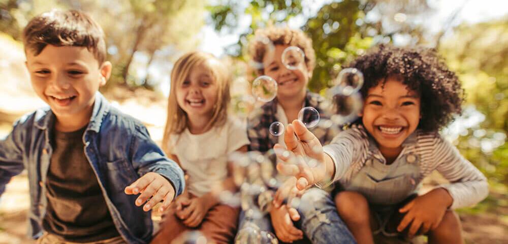 Vier lachende Kinder spielen fröhlich mit Seifenblasen im Freien an einem sonnigen Tag.