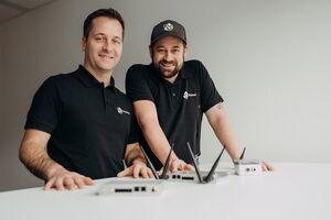 Two men in branded shirts smile while showcasing electronic devices on a white table.