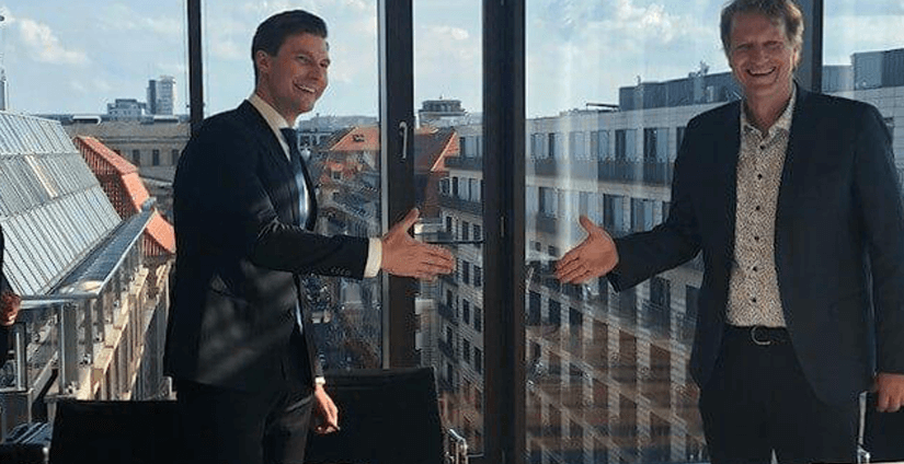 Zwei Männer in Anzügen stehen in einem modernen Büro vor einer Glasfront und bereiten sich auf einen Händedruck vor.