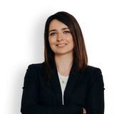 Eine Frau in lässigem Outfit lächelt selbstbewusst mit verschränkten Armen vor einem weißen Hintergrund.