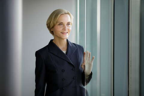 A confident woman in a dark blazer stands by a window, gazing thoughtfully outside.