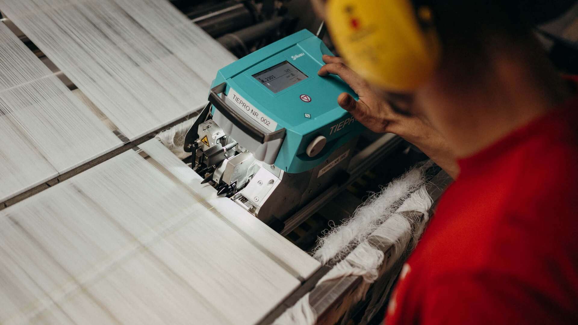 A worker in a red shirt operates a teal machine processing fabric on an industrial conveyor.