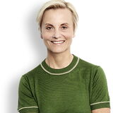 Eine Frau in einem grünen Oberteil steht lächelnd mit verschränkten Armen vor einem weißen Hintergrund.