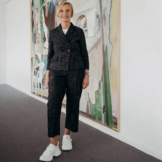 A smiling woman in a dark suit and white sneakers stands next to a colorful abstract painting in a bright room.