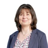 A woman in a patterned blouse and navy blazer smiles confidently against a plain white background.