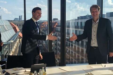 Two men in suits stand smiling, about to shake hands in a bright office with a city view.