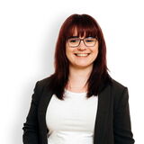 Eine lächelnde Frau mit Brille und schwarzem Blazer steht lässig vor einem weißen Hintergrund.