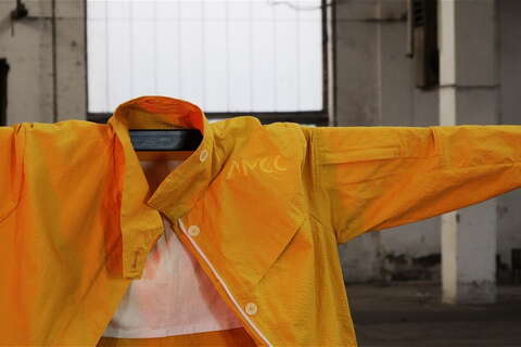 A vibrant orange jacket with "AMCC" displayed on the chest is spread open on a hanger in an industrial setting.