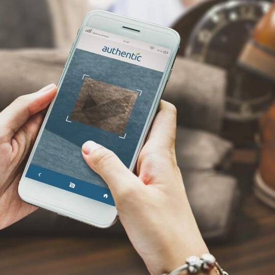authentic - BUSINESS AREAS - Smartphone A person uses a smartphone app to verify the authenticity of a fabric, with shoes and a clock in the background.