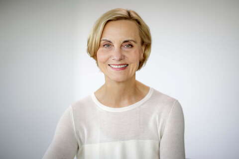 A smiling woman with short blonde hair is wearing a white blouse against a soft gray background.