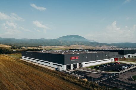 The ERTEX industrial building is surrounded by scenic hills, with parked cars in front and clear blue skies above.