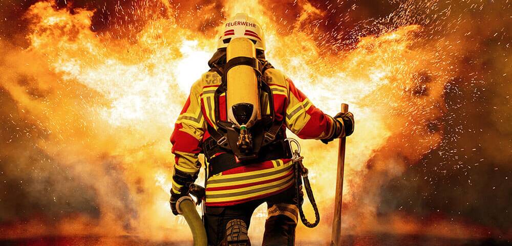 Ein Feuerwehrmann kniet mit Schlauch und Axt vor einem lodernden Feuer und bekämpft die Flammen mutig.