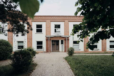 A brick building with a "Kontor" sign is framed by lush trees, creating a serene atmosphere.