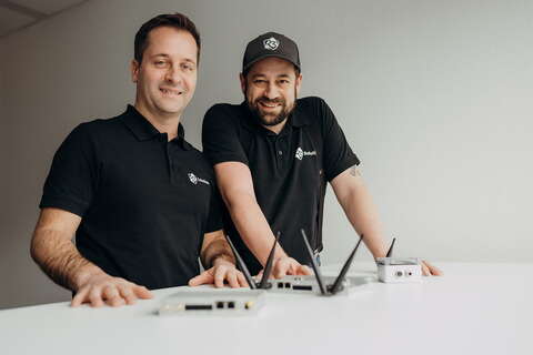 Two men in branded shirts smile while showcasing electronic devices on a white table.