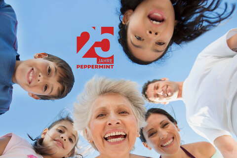 A joyful group of people of various ages smile down at the camera under a clear blue sky, celebrating 25 years of Peppermint.