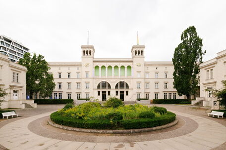 The elegant building features green accents and a circular garden, set against a cloudy sky. (Text KI-generiert)