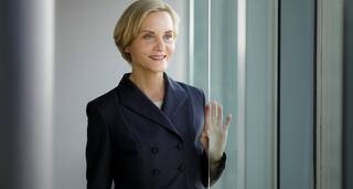 Eine Frau in einem eleganten Anzug blickt lächelnd aus einem großen Fenster.