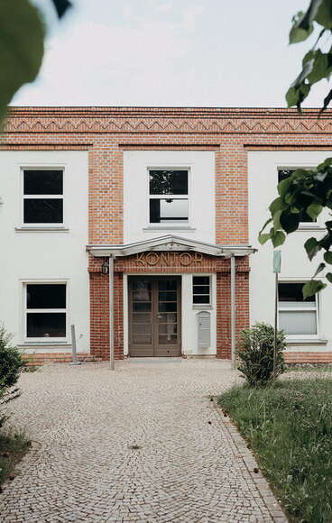 A brick building with a "Kontor" sign is framed by lush trees, creating a serene atmosphere.
