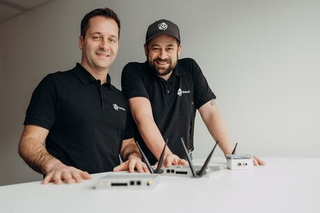 Zwei Männer in schwarzen Poloshirts stehen lächelnd an einem Tisch mit mehreren technischen Geräten.