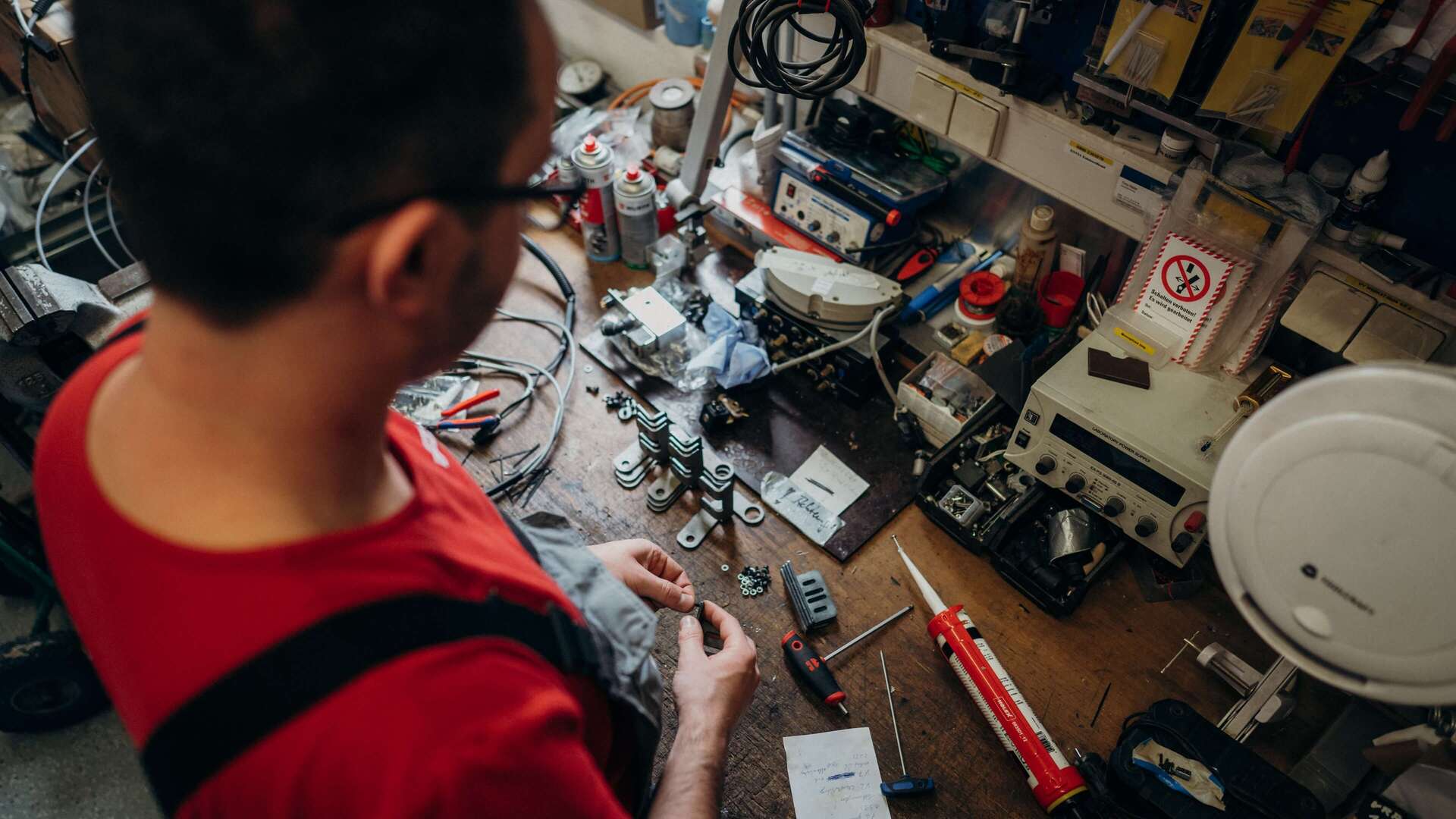 A person in a red shirt works on electronics at a cluttered workbench filled with tools and components.