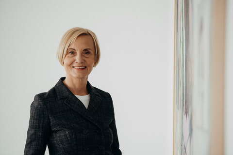 A smiling woman in a dark blazer stands near a large piece of art on a white wall.