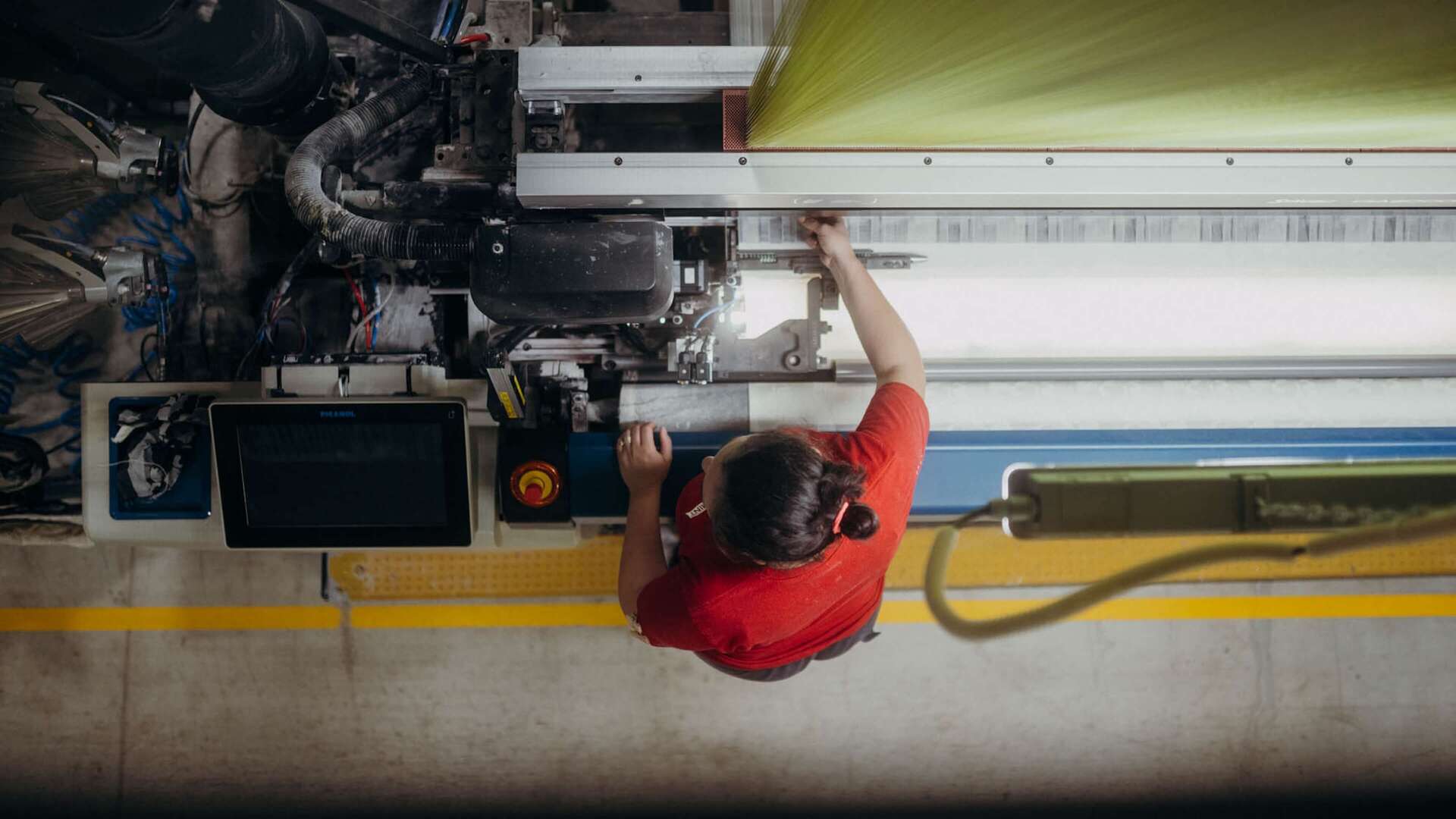 A person in a red shirt operates a complex industrial machine from above, adjusting controls with precision.