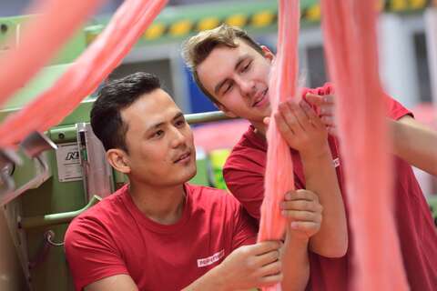 Two men in red shirts are carefully inspecting pink yarn in a textile factory.